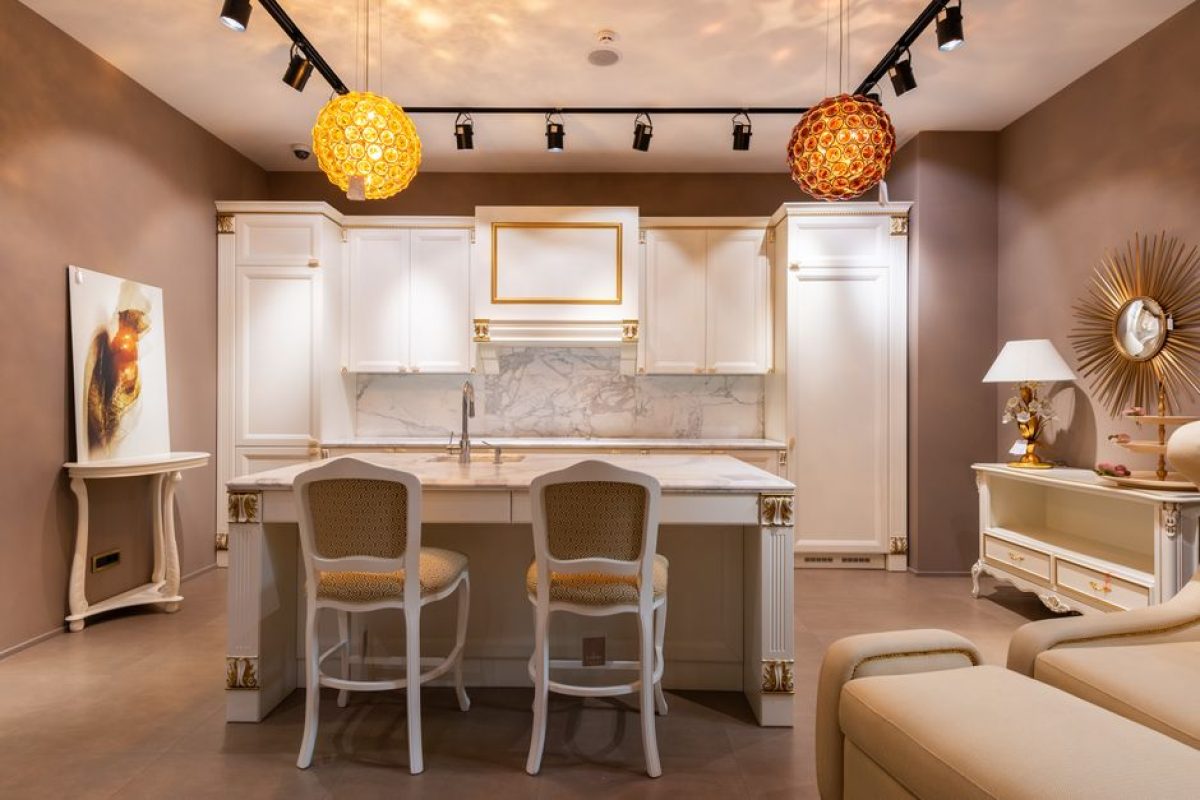Chairs at table placed near white cupboards in spacious kitchen with glowing lights and cabinet with lamp and decorations in apartment